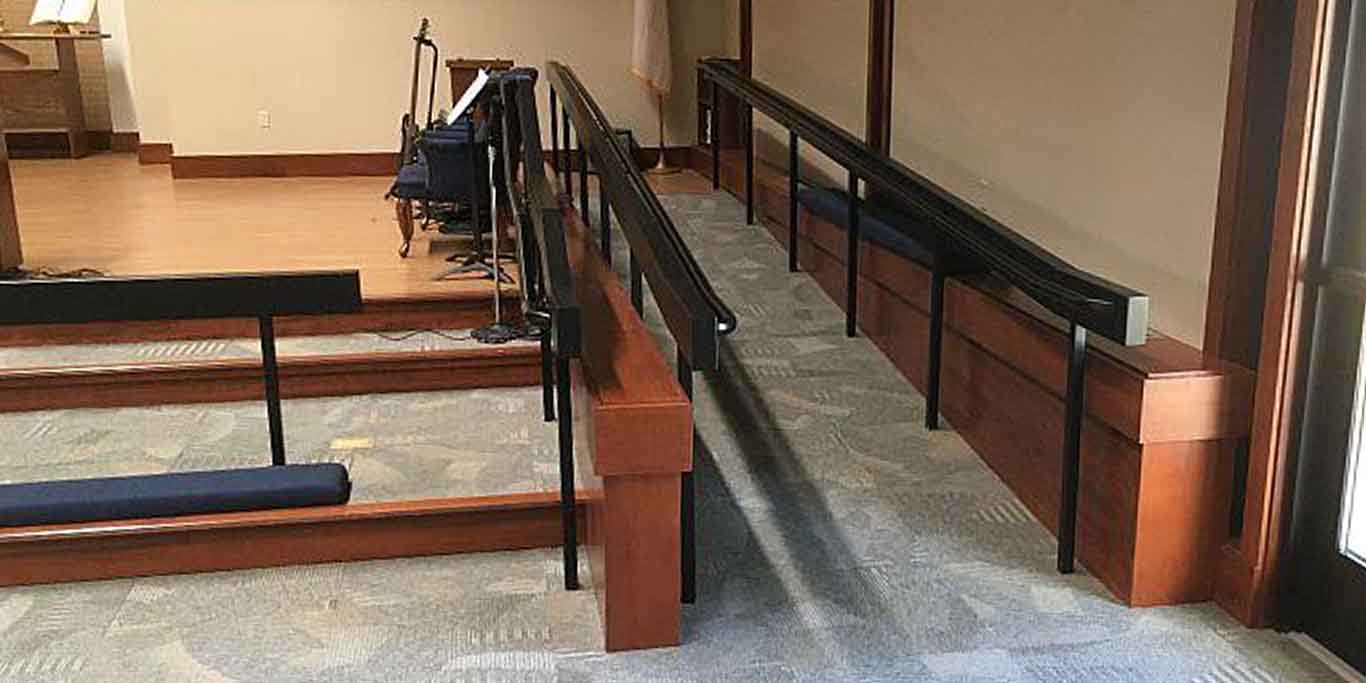 A picture of a wheelchair ramp on the side of a church chancel, providing access. A kneeling rail and steps are in place on the left side of the image and the ramp is along the wall to the right.