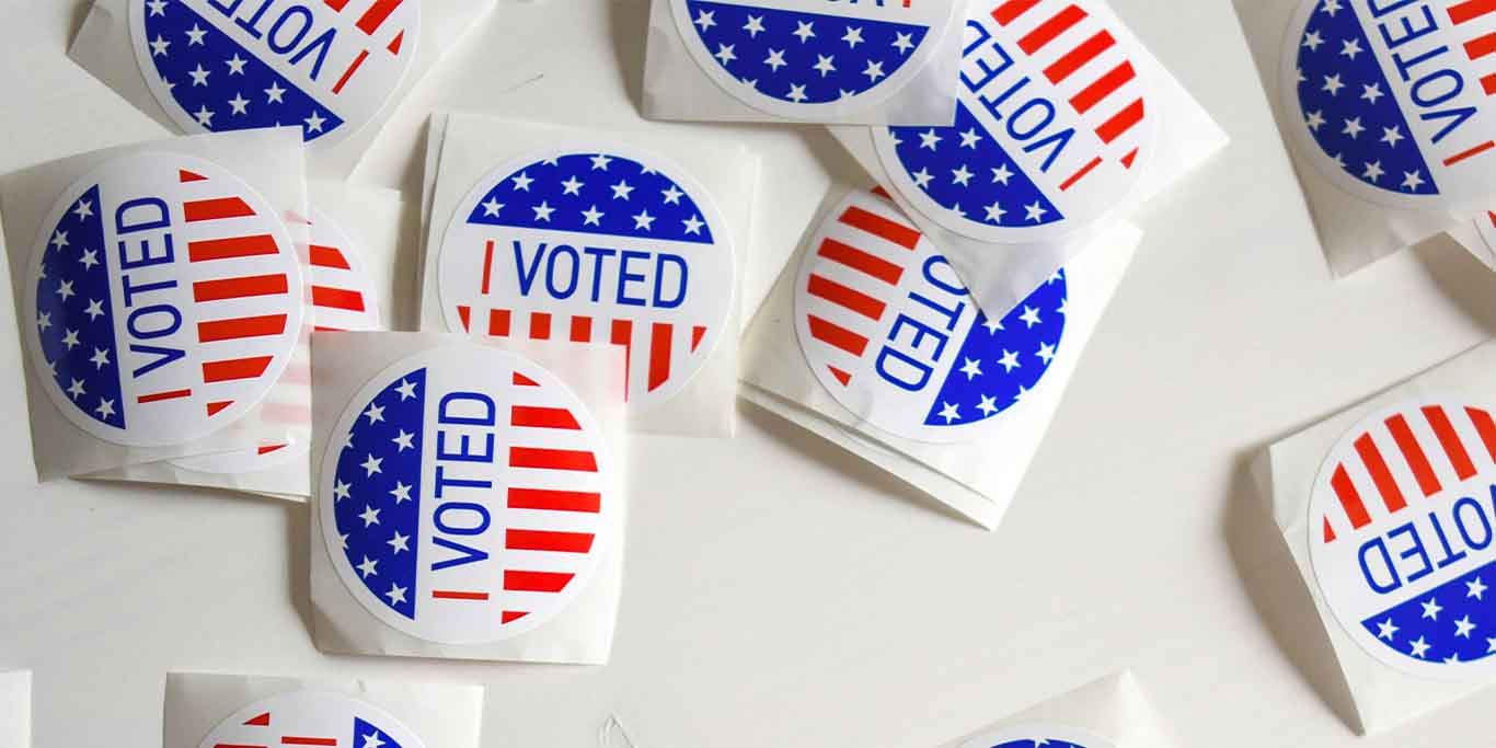 A bunch of stickers on a table with the words I VOTED. These are stickers you would commonly find at polling places. The stickers are circular, still attached to a square baking paper. The top third of the sticker contains white stars on a blue background. The middle of the sticker says I VOTED. The bottom third of the sticker features red and white strips running vertically.