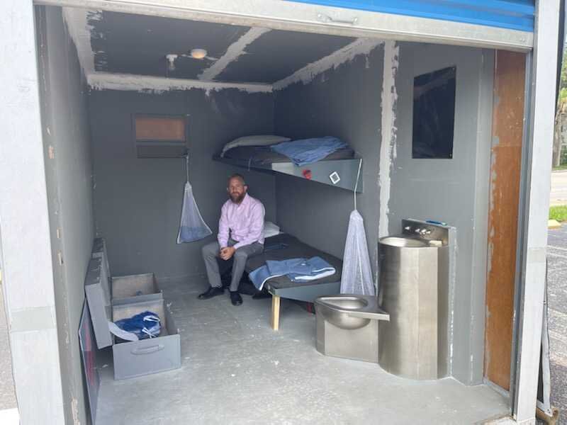 A man sits inside a replica of a Florida prison cell. He is wearing a pink shirt and grey pants and is sitting on the bottom bunk of a bunk bed attached to the wall.