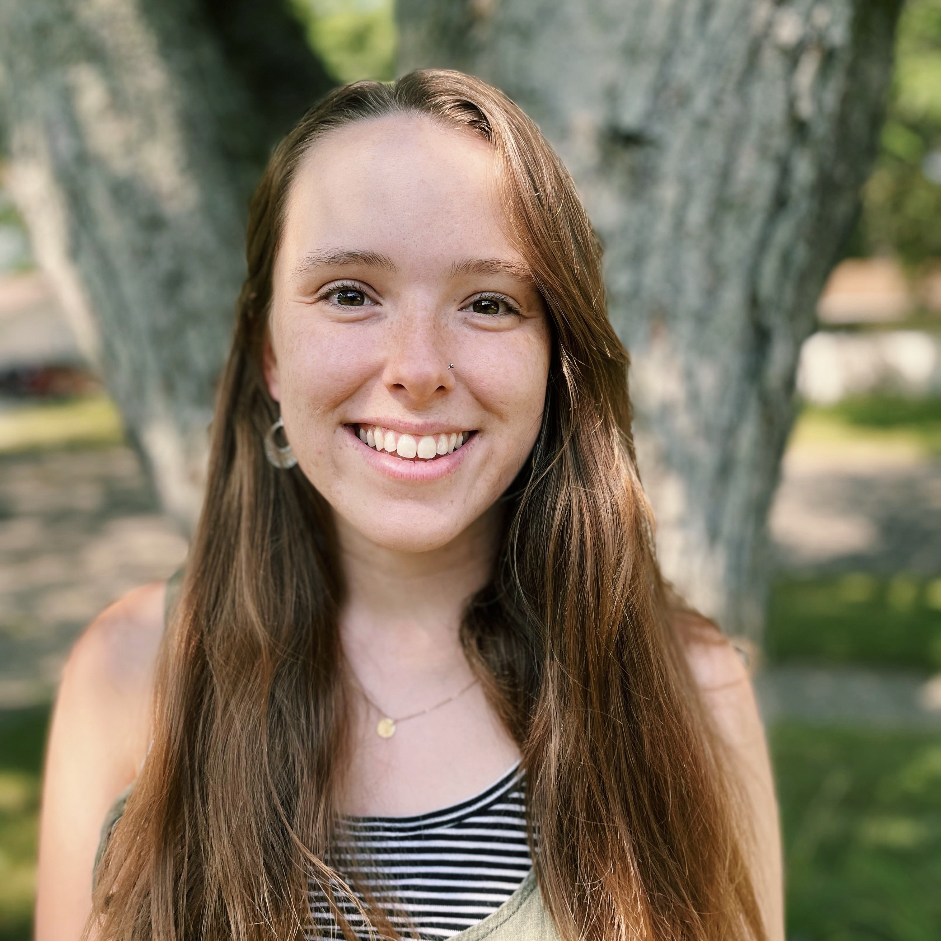 A photo of Emily smiling in front of a tree. She is wearing a striped shirt.