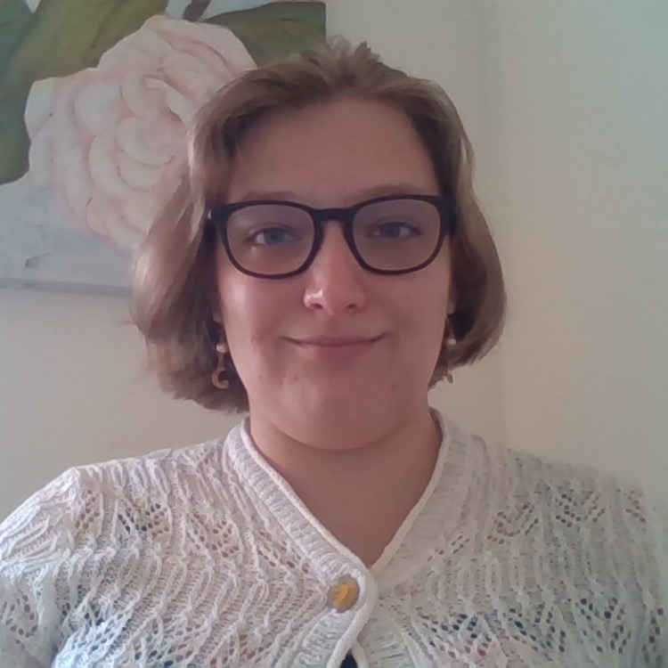 A photo of Sarah in a white sweater smiling sitting in front of a picture of a flower.